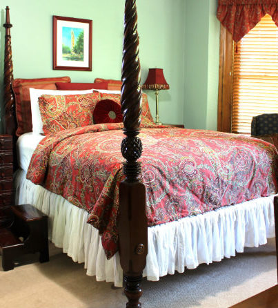 4 poster bed with red duvet and large pillows. Wingback chairs are visible in front of bay window